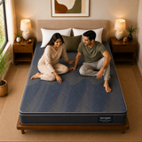 A man and a woman sit on a dark blue Springtek Euro Top Hybrid Latex Pocket Spring Mattress in a cozy, symmetrically styled bedroom with wooden nightstands, lamps, and a beige headboard.