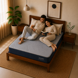 A couple relaxes on a Springtek Ortho Pocket Memory Foam Spring Mattress in a cozy, warm-toned bedroom. The mattress features a textured light blue top and a dark navy blue base, set on a wooden bed frame with matching bedside tables and a potted plant in the background