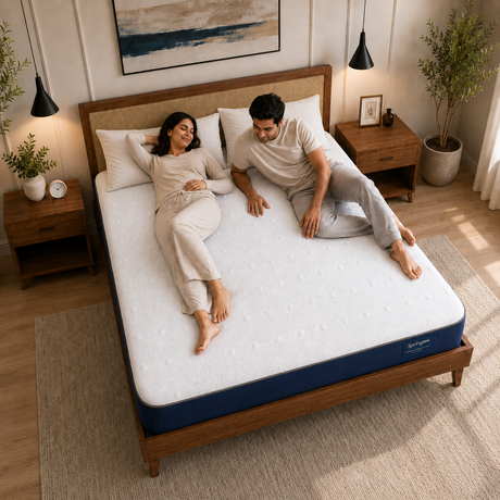 A couple relaxes on a white Wavemax Spine Support Orthopedic Mattress with a blue edge trim on a wooden bed frame in a warm, modern bedroom. The brand name 'Sensation' is visible on the side of the mattress.